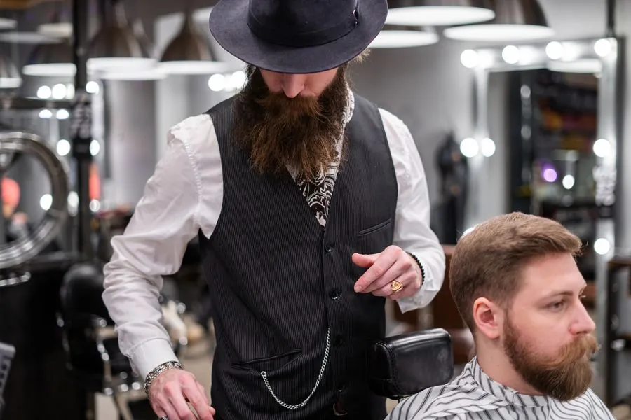 A barber in a waistcoat holding clippers beside a seated customer